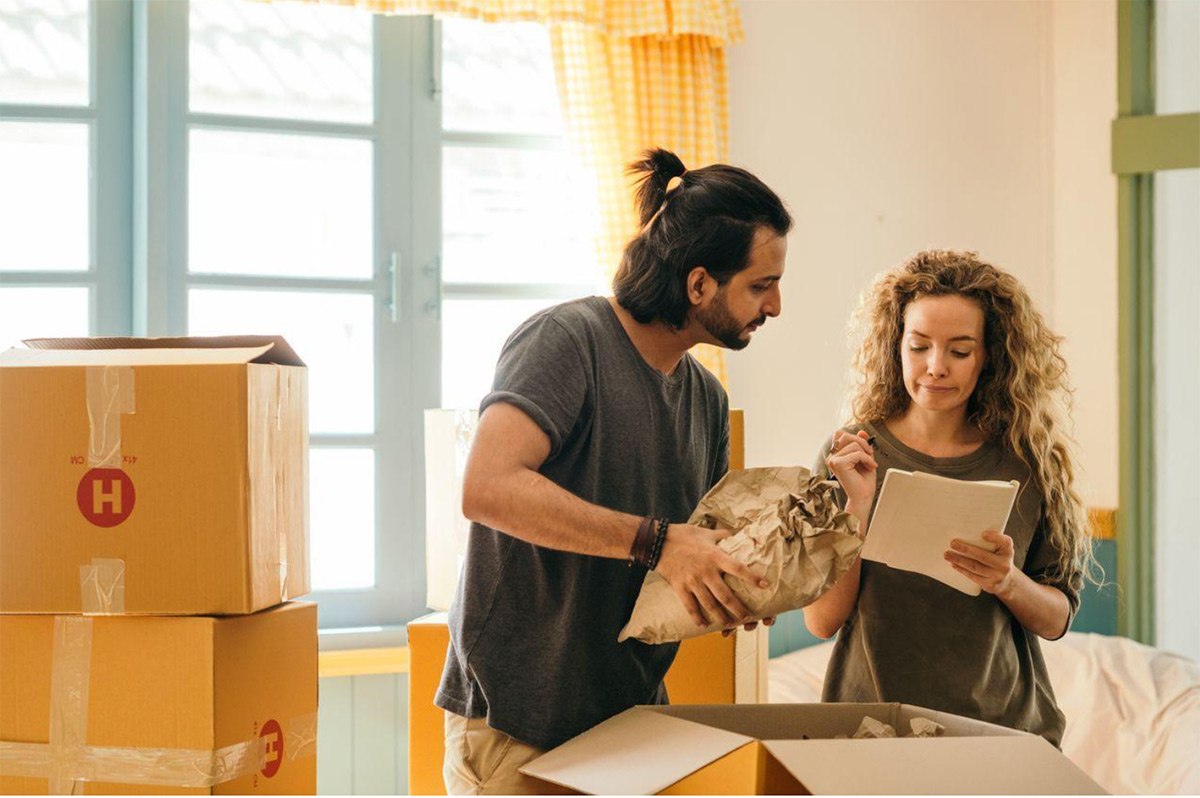 Newlywed-couple-unpacking-moving-boxes-together-while-checking-off-a-shared-home-checklist Newlywed couple unpacking moving boxes together while checking off a shared home checklist