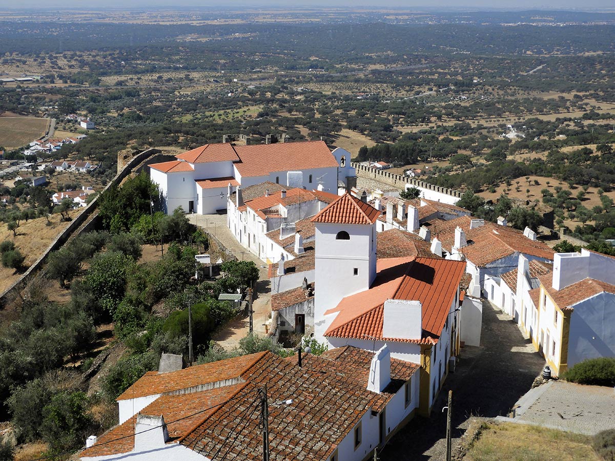 An elevated view over a whitewashed hilltop village with terracotta roofs and old fortress walls above the Alentejo plain